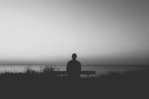 Man sitting alone on a bench, facing the sea under a grey evening sky, reflecting themes of grief and solitude