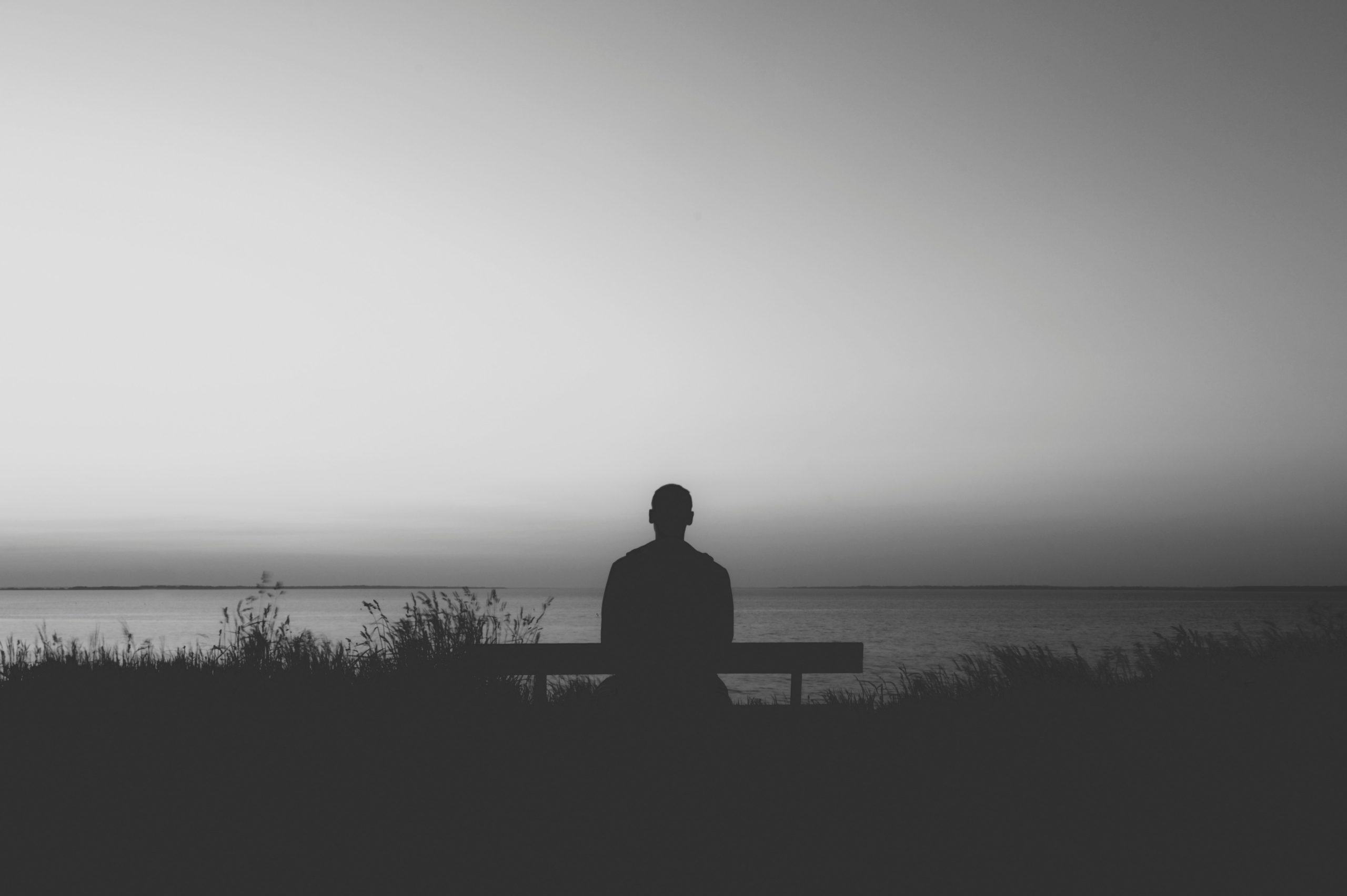 Man sitting alone on a bench, facing the sea under a grey evening sky, reflecting themes of grief and solitude